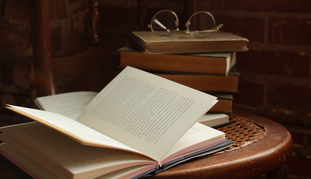 An open book mid flip with a stack of old books and a pair of glasses next to it